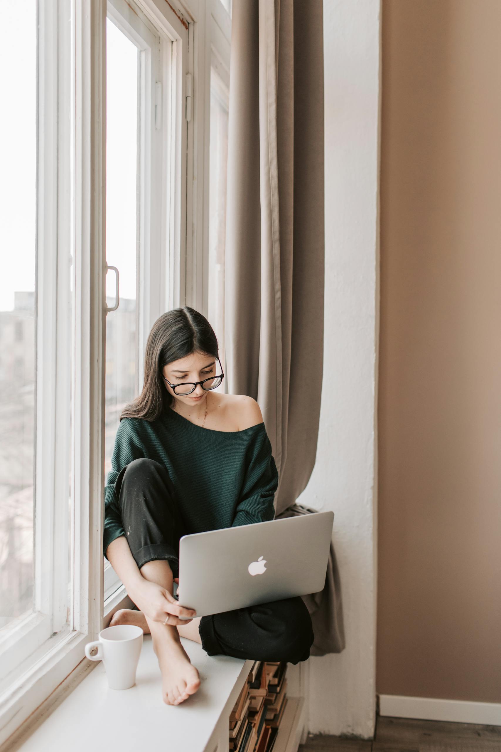 A relaxed young woman with glasses browsing her laptop while sitting on a windowsill during the day.